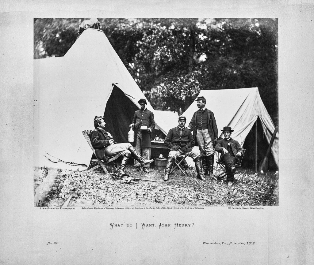 Three men are seated and two are standing in front of Civil War era tents. Four of the men are white, and the lone black man is delivering food to one of the men. The black man has a jug in one hand and a small plate in the other. Two of the five men are looking into the camera.