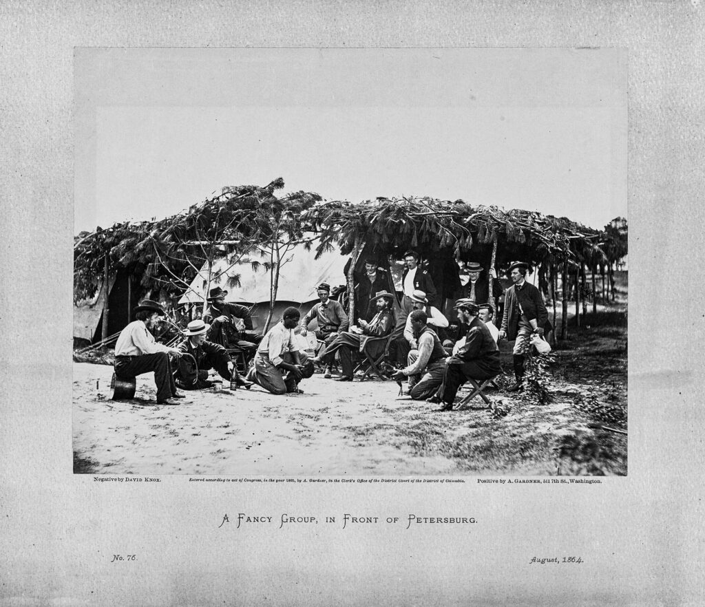 Twelve white men are sitting or standing, watching two black men facilitate a cockfight. Both black men hold birds in their hands while the twelve white men watch. The men are outside, on a Civil War battlefield, surrounded by makeshift housing and trees.