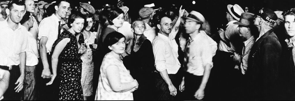 A large crowd of people assemble at the scene of a lynching. Many of the attendees are looking into the camera. Some look away. This image is a detail of the original photograph. The lynched bodies have been removed from the image.