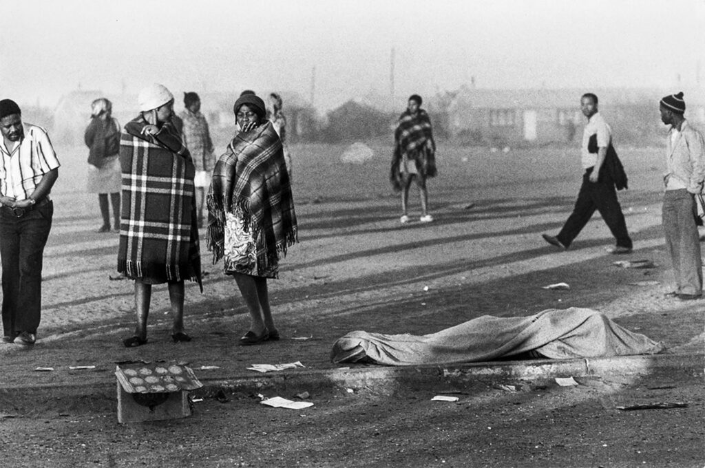 Nine people stand outdoors either looking at or disregarding a covered dead body in Soweto, South Africa.