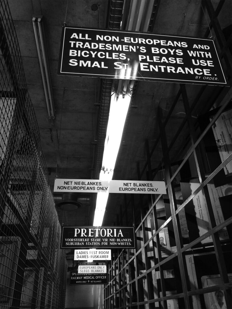 Interior of the Apartheid Museum in Johannesburg, South Africa. A South African apartheid era sign read’s “All Non-Europeans and Tradesmen’s boys with Bicycles, Please use Smal St. Entrance.” Signs further back read “Net Nie-Blankes/ Non-Europeans Only” and “Pretoria: Voorsteldelke Stasie Vir Nee-Blanks/ Suburban Station for Non-Whites.”