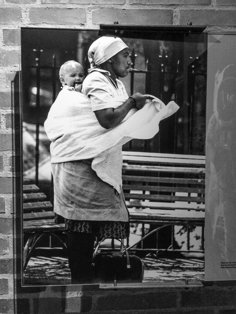 Exterior, Apartheid Museum, Johannesburg, South Africa. A photograph of a black woman in front of two benches. Her bag is on the floor to her left, and she is in the process of securing cloth around her waist with a white baby strapped to her back.