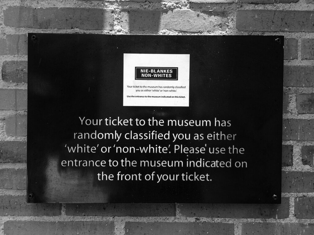 Exterior of the Apartheid Museum, Johannesburg, South Africa. A sign affixed to a brick wall states: “Your ticket to the museum has randomly classified you as either ‘white’ or “non-white.’ Please use the entrance to the museum indicated on the front of your ticket.”