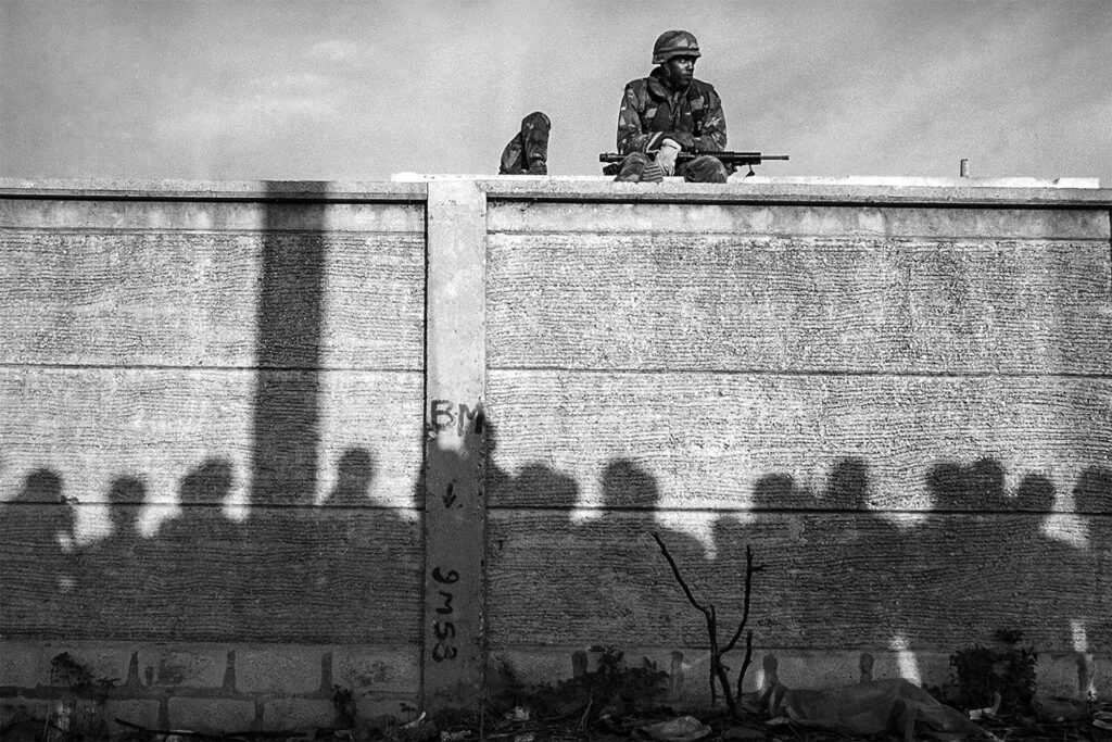 A U.S. soldier is seen sitting on top of a wall. He has an automatic rifle sitting across his lap. He is looking to his left. Next to him there is another soldier facing away from the camera. There are shadows set against the wall and the shadows are the silhouettes of a group of people standing in front of the wall.