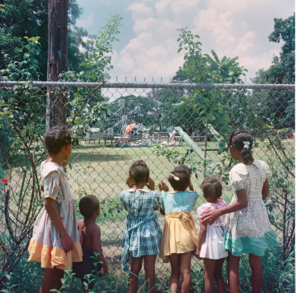 Six black children (five girls and one boy) stand against a fence outside a segregated playground with their backs to the viewer. The playground is in the distance in front of them.