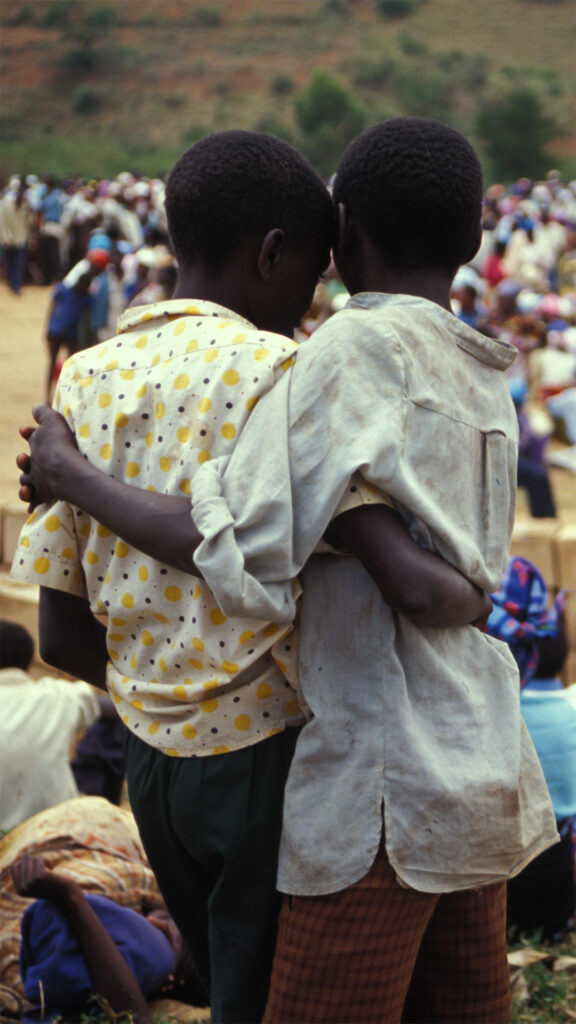 A photograph of two Rwandan children with their arms wrapped around each other. Their backs are facing the viewer. The children are in front of a large crowd of people.