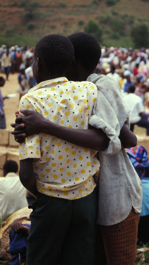 A photograph of two Rwandan children. One child has hands clasped around the other in a hug. The children are in front of a large crowd of people.